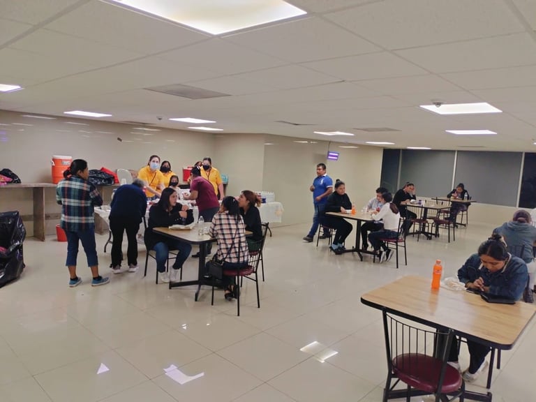 Group of diverse people in casual clothing gathered in a bright indoor room with tables and chairs during what appears to be a community or team activity